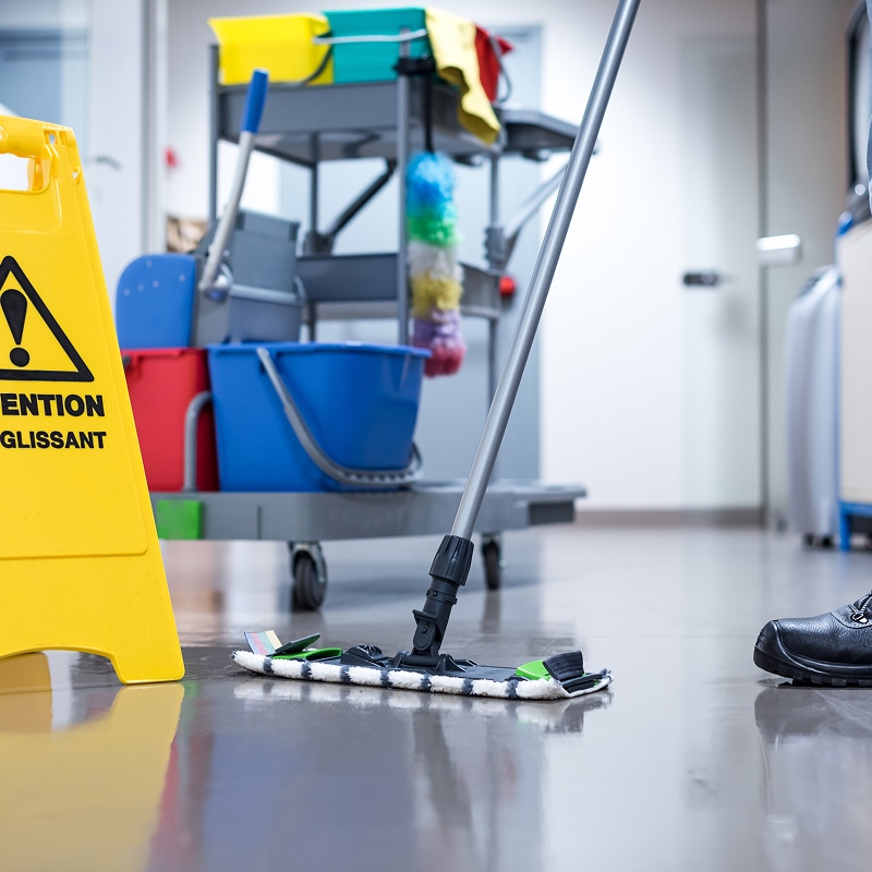 Sol brillant d'un couloir nettoyé, avec un balai-serpillière, un panneau jaune "ATTENTION GLISSANT" et un chariot de ménage en arrière-plan.