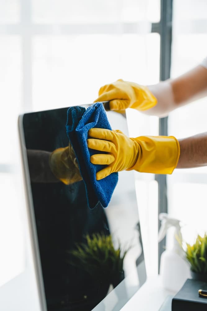 Nettoyage écran : Comment désinfecter votre ordinateur de bureau. Mains en gants jaunes essuyant l'écran d'un ordinateur de bureau avec une lingette bleue. Nettoyage et hygiène du matériel informatique.